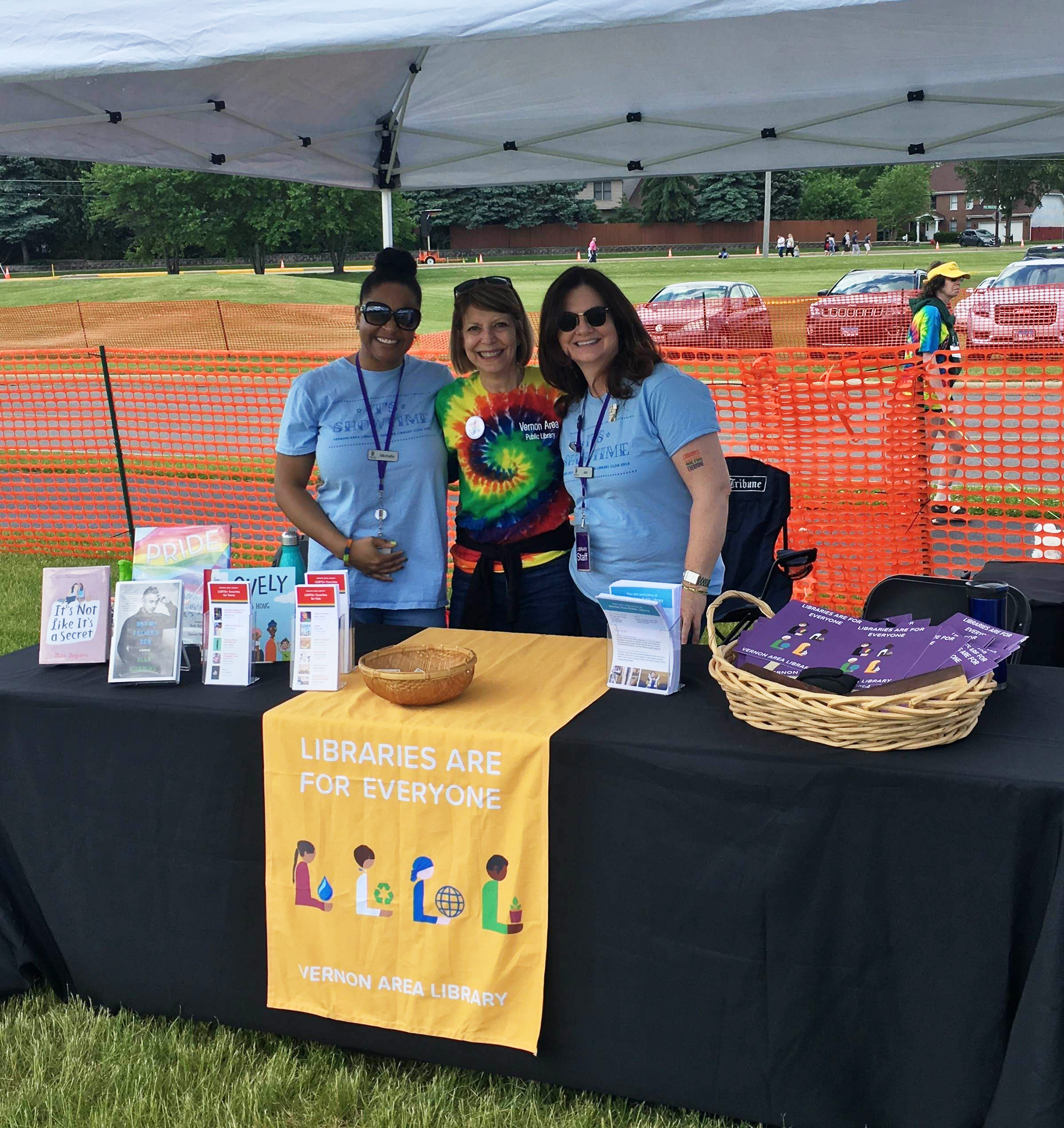 Photo of library Pride booth from 2019. A 6 foot table under a white tent awning is covered with rainbow-colored handouts. A banner on the table reads "Libraries Are For Everyone". Three smiling adult women stand behind the table. They wear library t-shirts.