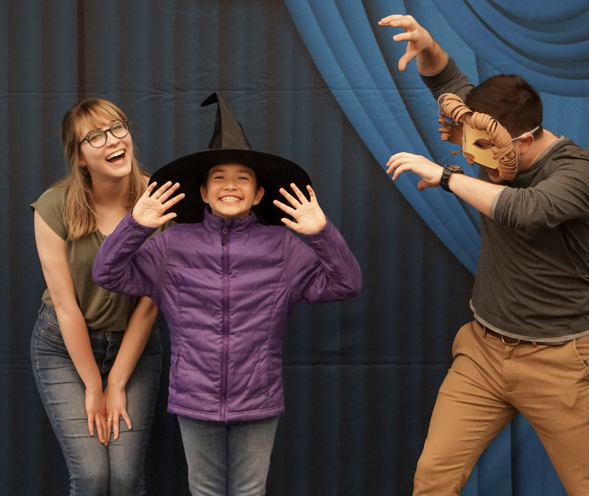 A young girl in street clothes and a witch hat makes a theatrical gesture. To her left, an adult woman bends down to her level and grins at the camera. To her right, an adult in a lion mask gestures toward her.