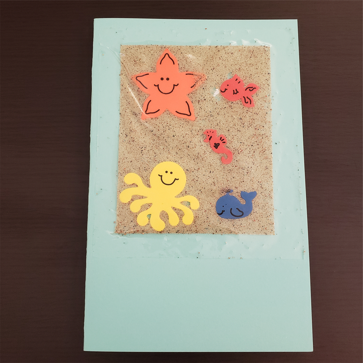 A beach greeting card: a rectangular folded paper, decorated with sandpaper and cutout shapes of sea creatures