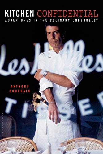 Anthony Bourdain is standing in front of a restaurant table wearing a chef's coat and apron.