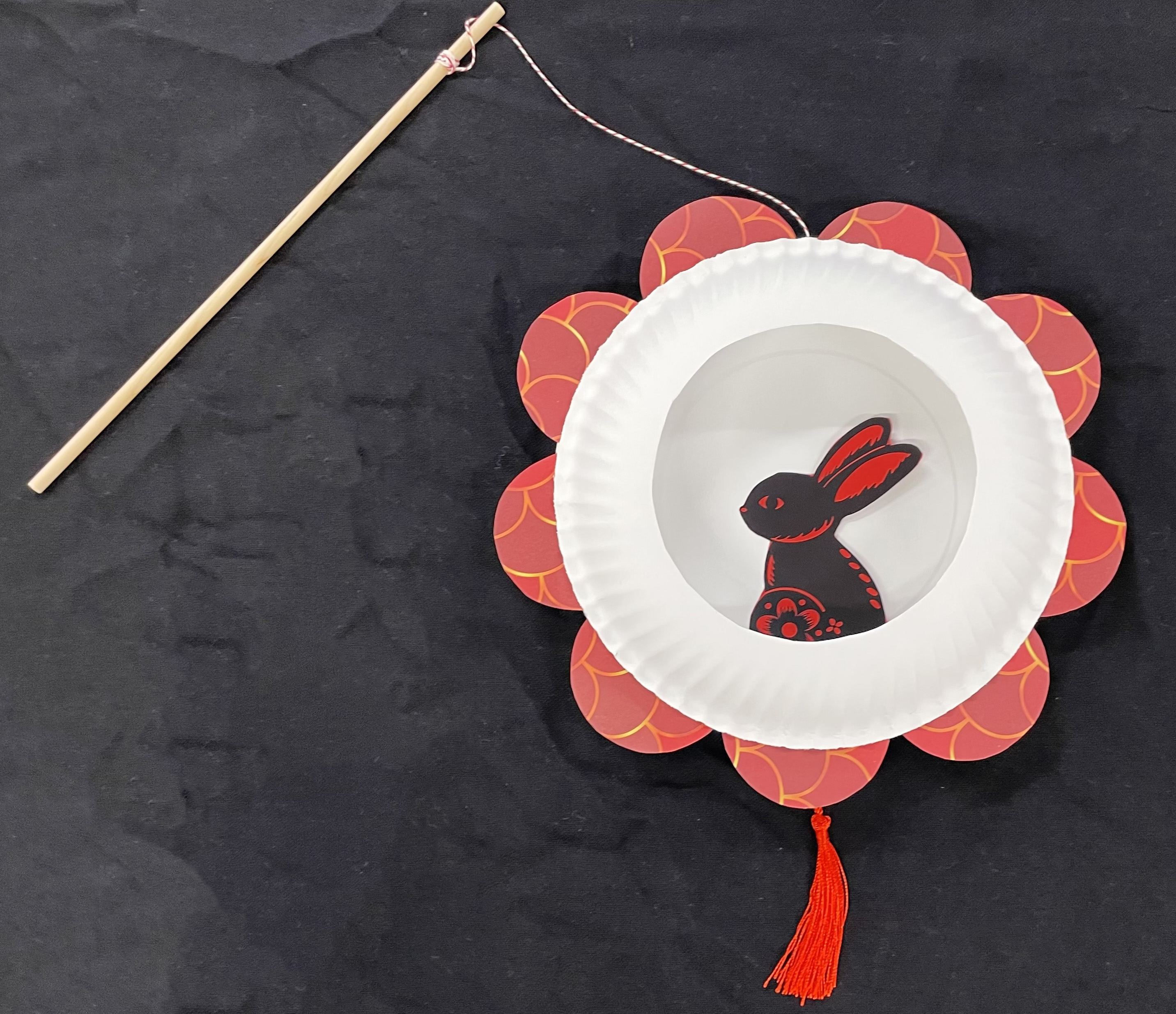 A paper rabbit inside a paper plate frame, decorated with red petals and a red tassle