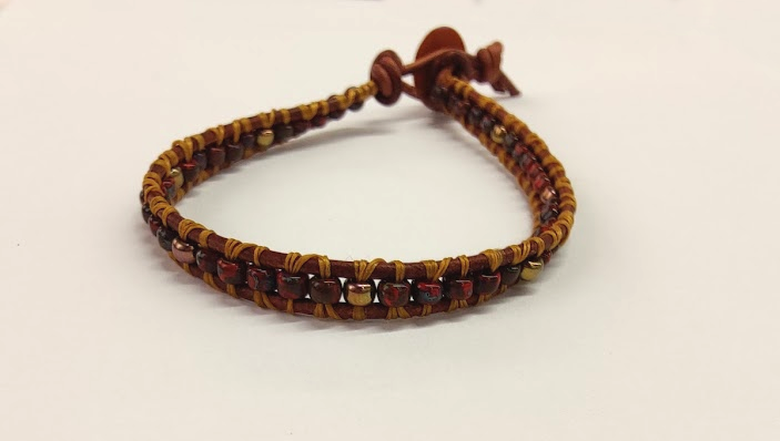 A beaded bracelet on a white background. The bracelet has brown leather cord, red and gold beads, and gold thread.