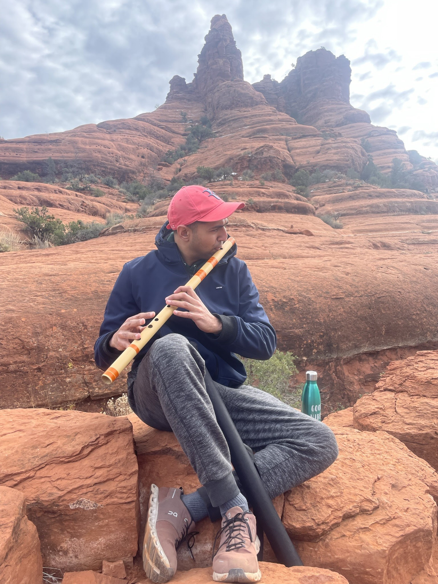 Man in blue sweatshirt, gray pants and red baseball hat sitting on red rock playing a wooden flute with mountains in the background.