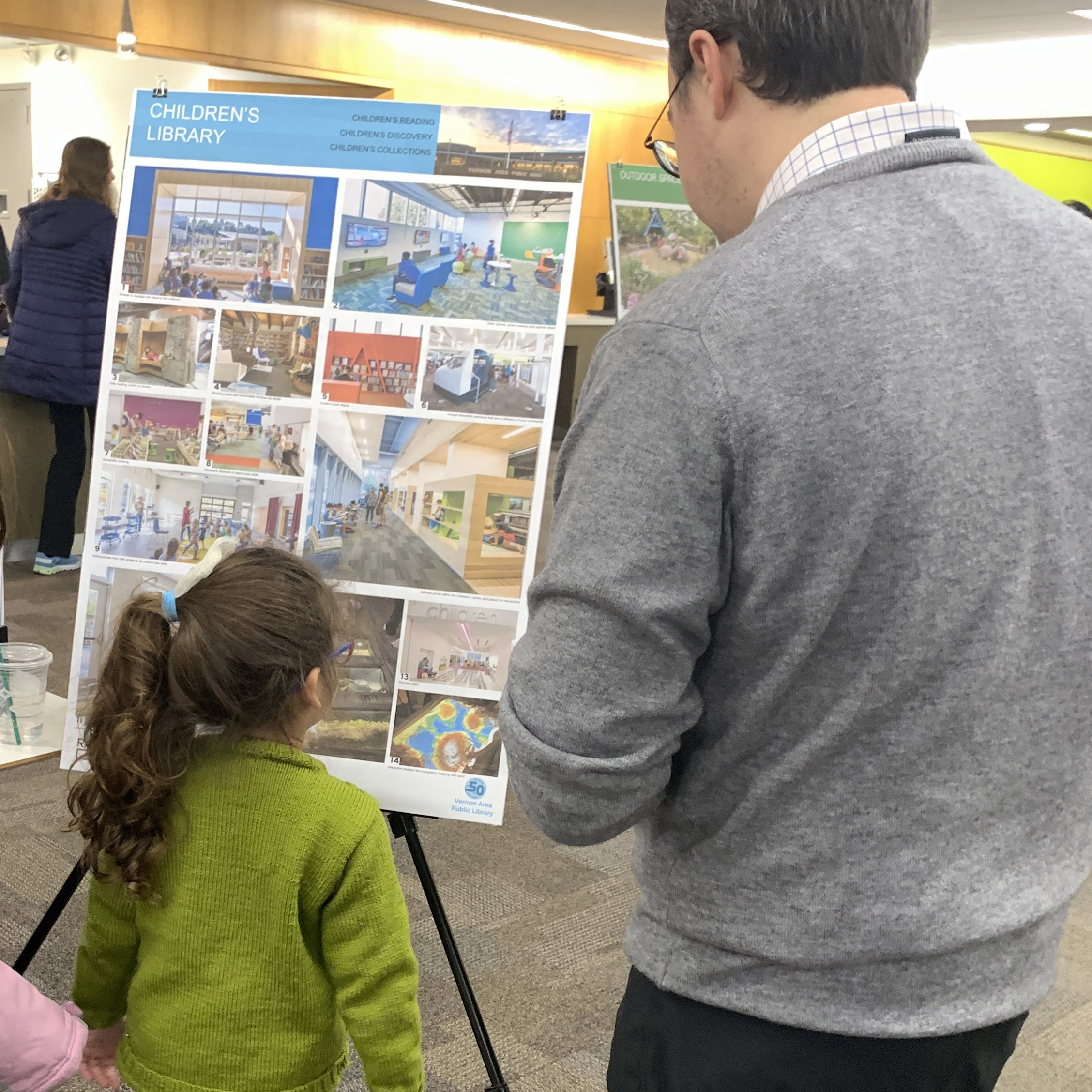 A young girl looks at a poster board filled with pictures of children's libraries. An adult man looks over her shoulder.