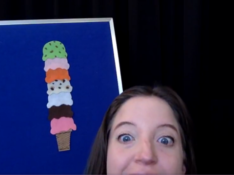 An adult woman with wide eyes peeks into the camera from the bottom of the frame. Behind her is a felt board with an ice cream cone of many colors.