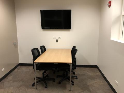 Photo of Adult Study Room 1 showing a table with 4 rolling office chairs. On the wall is a large TV screen. Some input sockets are seen beneath.