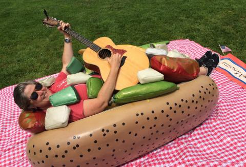Musician Laura Doherty in an outdoor setting. She lies on an inflatable hot dog bun as if she was the sausage. She wears sunglasses and plays a guitar.