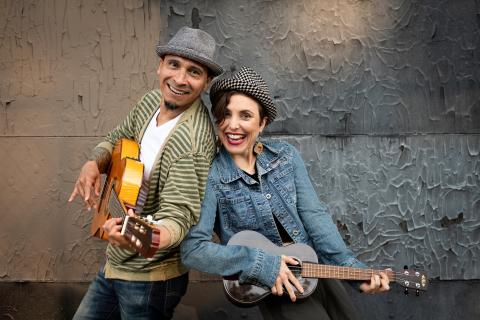 A Hispanic man and woman stand back-to-back, grinning and holding guitars. 