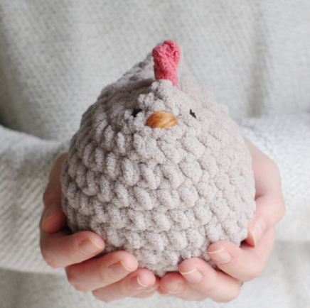 Plush gray chicken with a yellow beak and red comb, held in two hands against a light gray background.