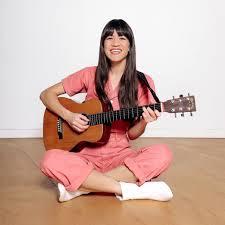 Woman with long dark hair in a pink jumpsuit sitting cross legged on the ground holding a guitar.