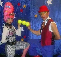 Woman in silver jumpsuit and pink hair and man in red vest and hat holding tennis balls.