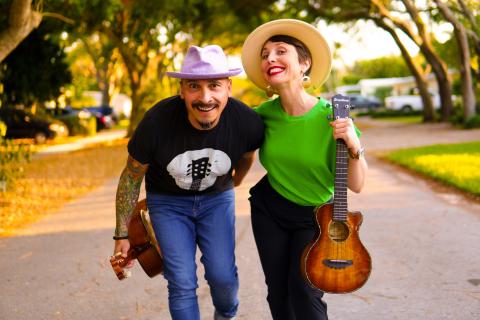 Man in hat and black t-shirt holding guitar walking arm in arm with woman in hat and green shirt holding ukulele