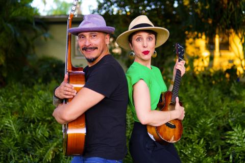 A man in a fedora and blue shirt hold a guitar back to back with a woman in a fedora and green shirt holding a ukulele.