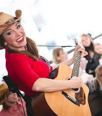 Woman with blond hair and freckles in a cowboy hat and red shirt holding a guitar.