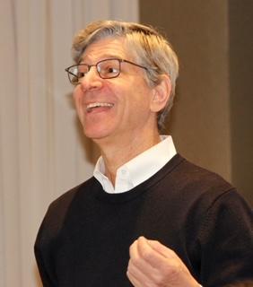 Man with gray hair and glasses wearing a white shirt and brown sweater