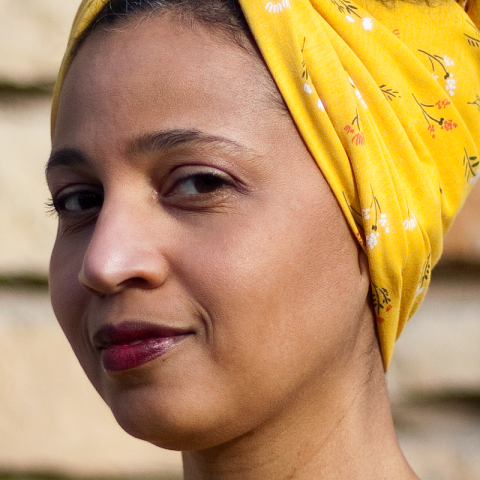 Headshot of a Black woman with a yellow headscarf and a wry expression