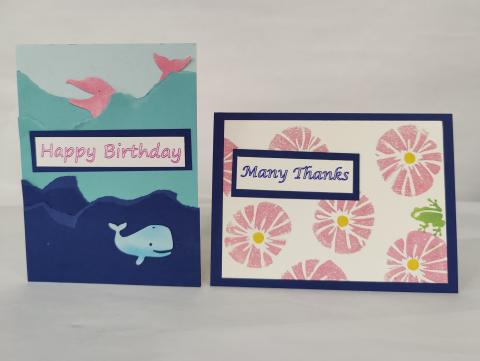 Two handmade cards, one with an under-the-sea theme with a whale and dolpih that says "Happy Birthday." Another with pink and yellow stamped flowers that says "Many Thanks"