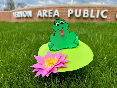 Green paper frog on a light green paper lily pad with a pink flower sitting on the green grass in front of the brink wall with the Vernon Area Public Library sign.