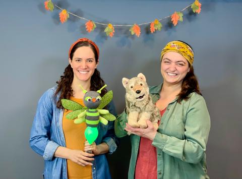 Two women with brown hair and long sleeve shirts holding a wolf puppet and plant
