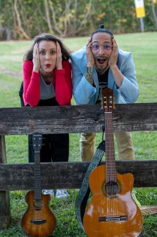 Woman with long brown hair and red lipstick with her elbows on a fence cradling her cheeks next to a man with dark hair and a light denim shirt with his elbows on the fence cradling his cheeks. a guitar and ukulele 