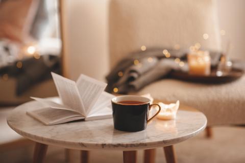 There is a table with an open book on it, a mug of coffee, and a candle. In the background, there is a chair with a blanket, a candle, and string lights. 