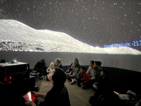 Adults and children sitting on the floor under the night sky projected onto the walls and ceiling