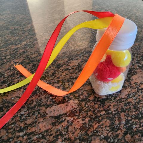 Small clear bottle holds rice with red and yellow pom poms inside. On top are three ribbons: orange, yellow, and red