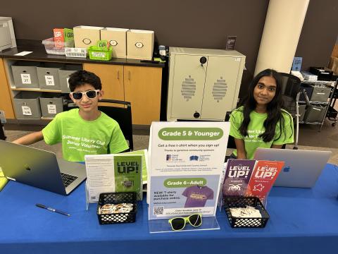 Teen volunteers wearing lime green shirts sit at a table in the library ready to sign people up for Summer Library Club