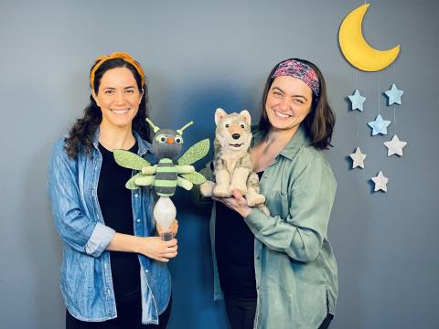 Two women with long brown hair wearing denim shirts and black pants holding a firefly and a wolf puppet