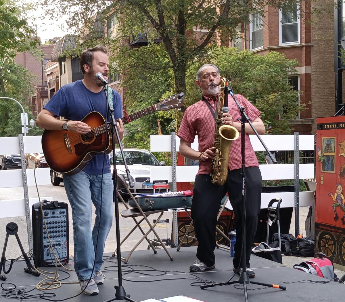 An adult man playing a guitar and an adult man playing a saxophone on an outdoor stage.