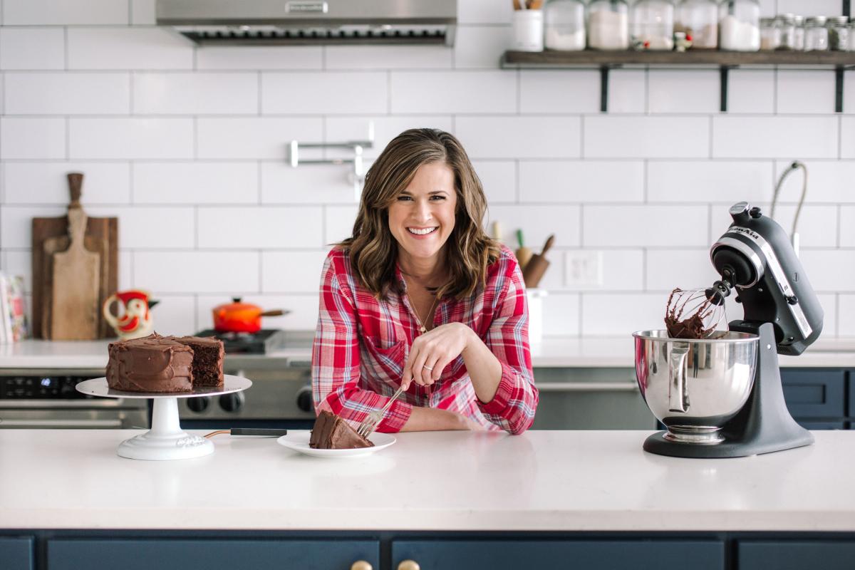 Baker Shauna Sever is a young woman with should-length brown hair. She leans on a kitchen counter. On the counter is a cake and a stand mixer.