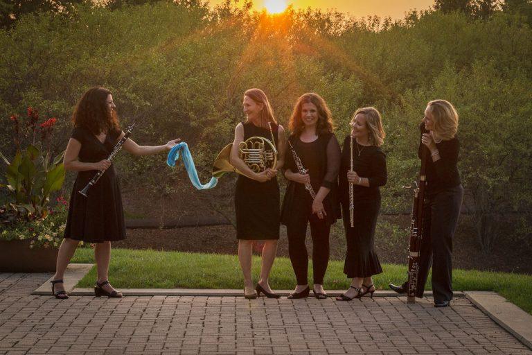 Four women in black dresses holding woodwind instruments while standing on a brick patio with trees in the background.  