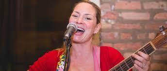 Woman with blond hair pulled back in a ponytail wearing a red sweater singing into a microphone and holding a guitar