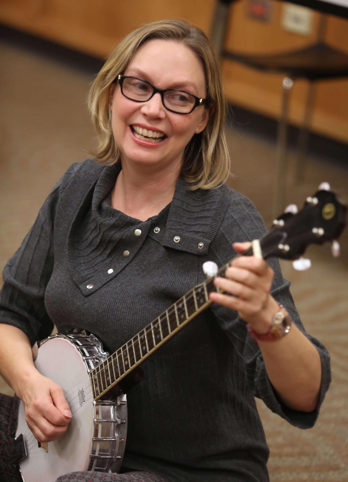 Woman with blond hair and glasses in gray shirt playing guitar