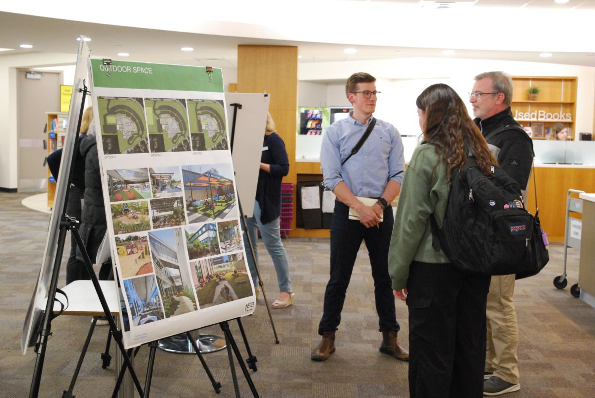 Two adults in fall coats chat with an architect who stands next to a vision board on an easel.