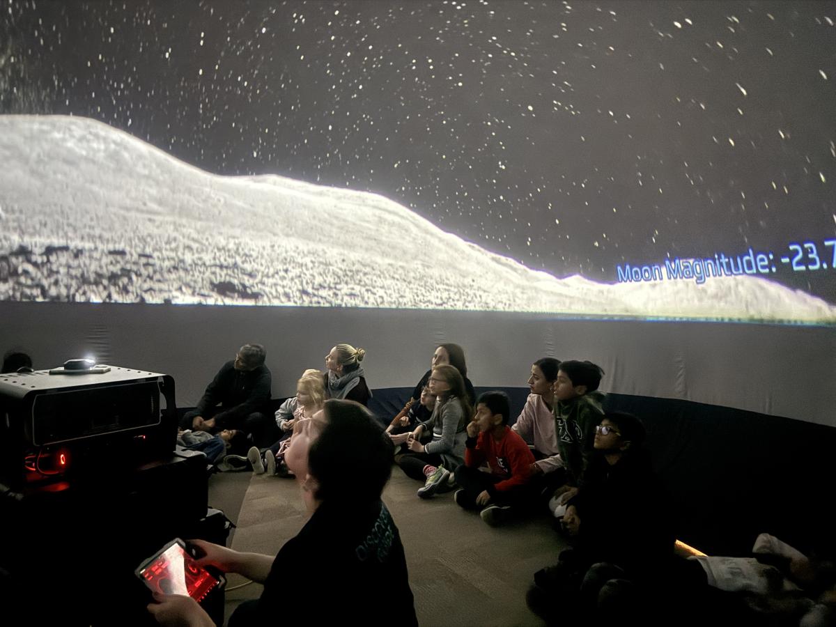 Adults and children sitting on the floor under the night sky projected onto the walls and ceiling