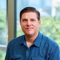 A man with short brown hair wearing a blue shirt in front of a window 