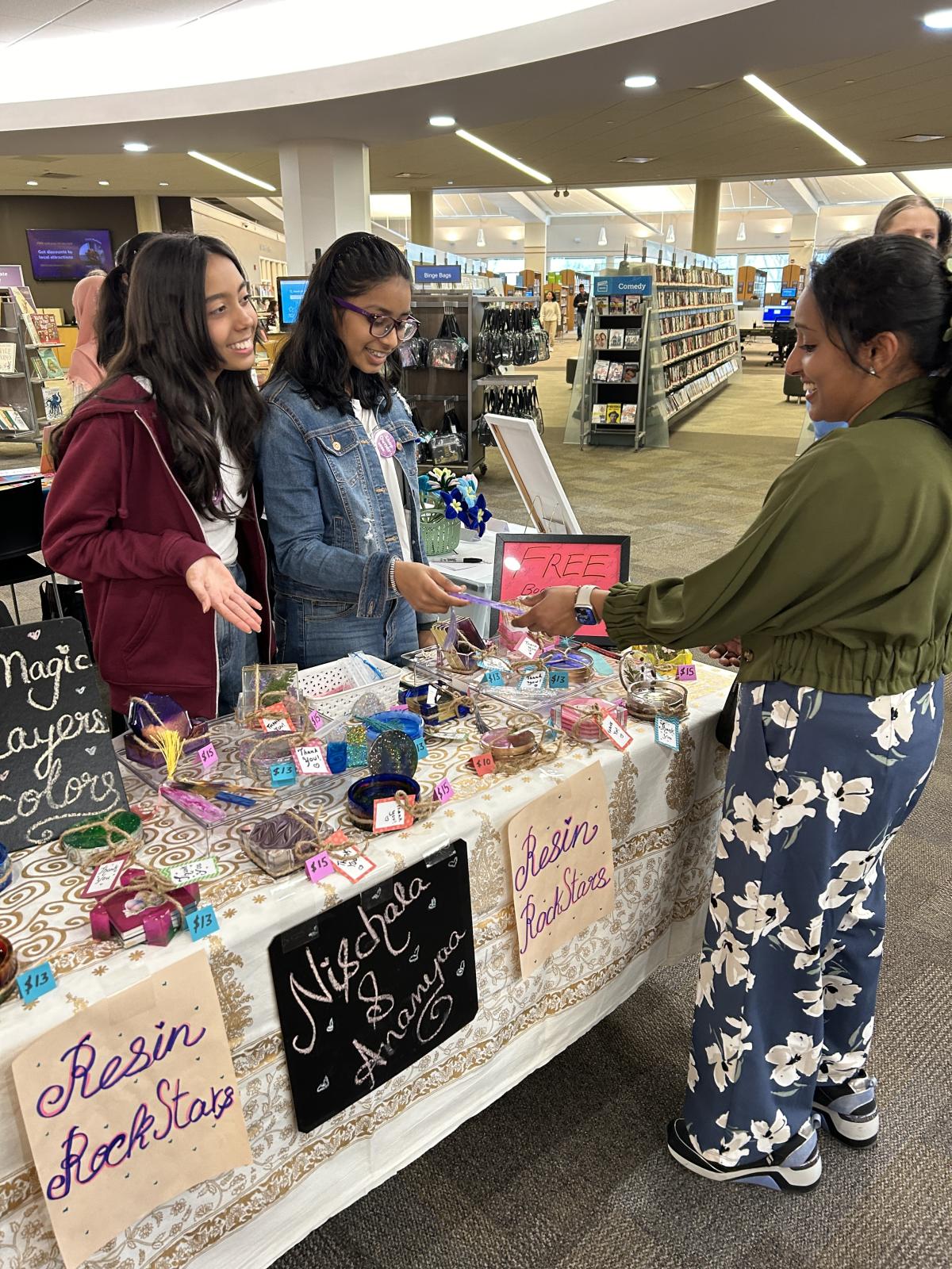 Two students interact with a customer at a sales table with homemade goods