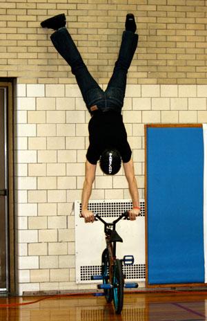 Man in black tshirt and jeans wearing a black helmet doing a handstand on a bike handle bars
